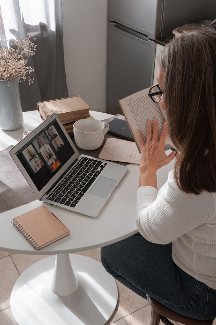 A woman participates in an online book club meeting using her laptop while holding a book at a home table.