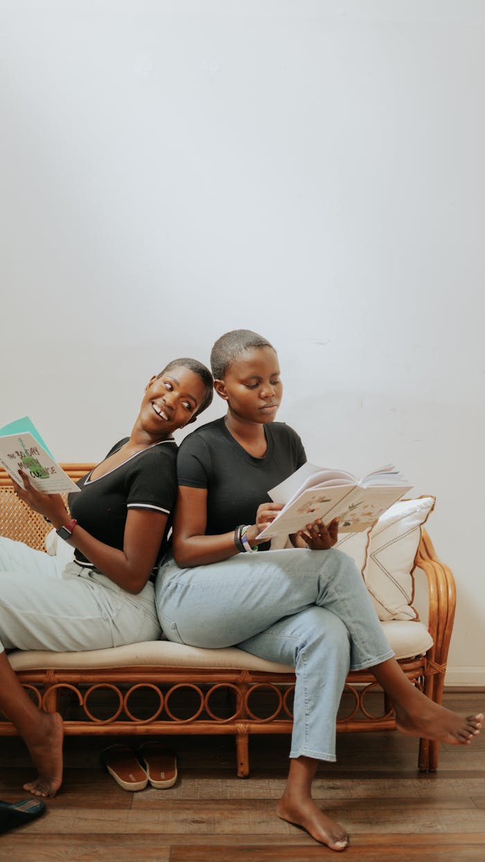 Two women sitting back-to-back on a rattan sofa, enjoying books in a cozy indoor setting.